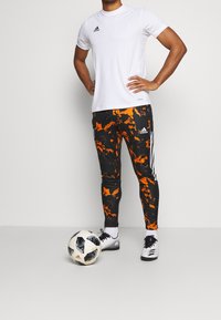 Bílý sportovní tričko s černým logem, spárované s černými legínami s oranžovými abstraktními vzory, vedle fotbalového míče a kopaček.