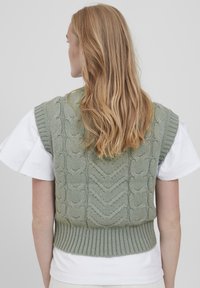 Strick-Westenpullover in sanftem Grün mit einem Zopfmuster, geripptem Saum und kurzen Ärmeln über einem weißen Hemd. Strukturiertes, tailliertes Design.