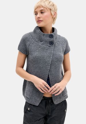 Vrouw draagt een grijze, gebreide korte mouw cardigan met twee grote knopen bij de hoge kraag, gecombineerd met een zwarte broek.