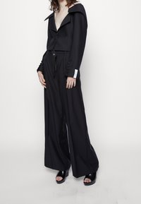 Svart skräddarsydd jumpsuit med off-the-shoulder-design, vida ben och knappar på ärmarna. Slät tygstruktur.