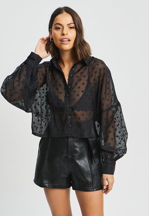 Blusa nera trasparente con pois, chiusura frontale con bottoni e maniche a palloncino. Abbinata a pantaloni corti in pelle nera a vita alta.