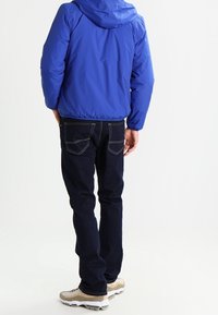 Blaue Kapuzenjacke mit elastischen Bündchen, getragen über dunkelblauen Jeans. Schuhe sind beige mit weißen und grauen Akzenten. Rückenansicht, einfarbiger Hintergrund.