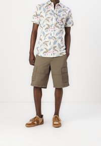 Camisa de manga corta con botones, con un estampado multicolor de hojas tropicales y flores, combinada con pantalones cortos cargo color caqui y zapatillas marrones.