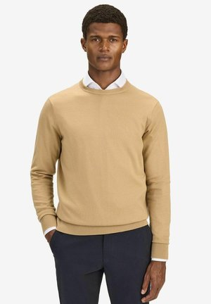 Beige strikket sweater med tætsiddende design, rund halsudskæring og ribbet kant. Båret over en hvid skjorte med krave, parret med mørkeblå bukser.