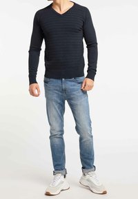 Muž v černém žebrovaném svetru s V-výstřihem, v světle modrých zúžených džínách a bílých teniskách stojící na bílém pozadí.