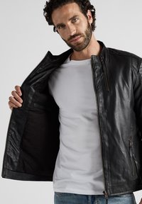 Zwarte leren jas met een ritssluiting aan de voorkant, staande kraag en ribbelstructuur. Draag open over een wit T-shirt, met een gladde binnenkant.