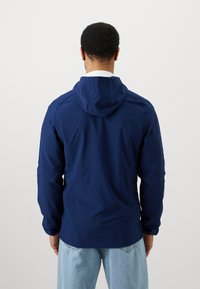 Casaco azul-marinho com fecho éclair e capuz, apresentando mangas longas e uma textura lisa e leve. A vista traseira destaca os detalhes das costuras.