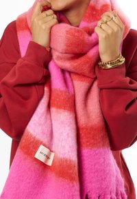 Scarf a strisce rosa e rosse realizzata in materiale morbido e peloso, con un'etichetta con scritto "MYJEWELRY" e bordi frangiati per un ulteriore effetto tattile.