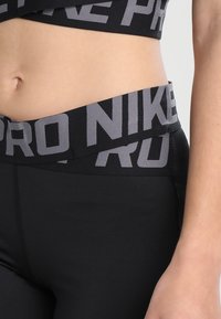 Leggings de sport noirs avec une large taille qui présente les marques "Pro" et "Nike" en gris. La texture lisse et la coupe ajustée améliorent le confort.
