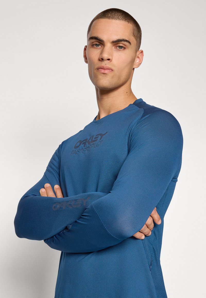 Camiseta deportiva de manga larga azul con un tejido texturizado, que presenta el logo "Oakley" en la parte delantera y en las mangas. Diseño de corte ajustado con cuello redondeado.