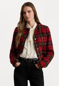 Sarkanais un melnais šalli auduma džemperis, pieguļošs dizains, apaļa apkakle, zelta pogas, uzvilkts virs baltas plandošas blūzes un melnām biksēm.