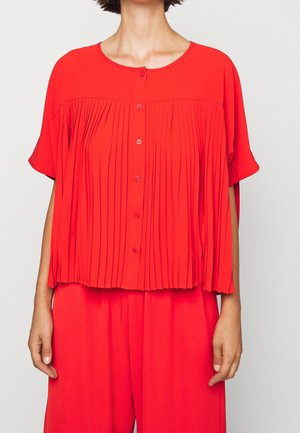 Femme portant un chemisier plissé rouge vif boutonné à manches courtes et un pantalon large rouge assorti.