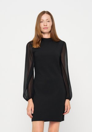 Femme aux cheveux bruns et raides, portant une robe noire à manches longues avec des manches transparentes, debout devant un fond blanc simple.