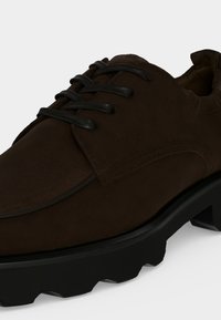 Chaussure à lacets en daim brun foncé avec une semelle épaisse en caoutchouc noir et une bande de roulement texturée, vue en gros plan sous un angle.