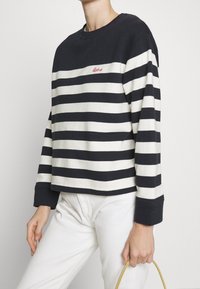 Námořnická a krémově pruhovaná mikina s dlouhým rukávem a kulatým výstřihem, s malým vyšívaným červeným logem na hrudi. Bílé kalhoty a taška.