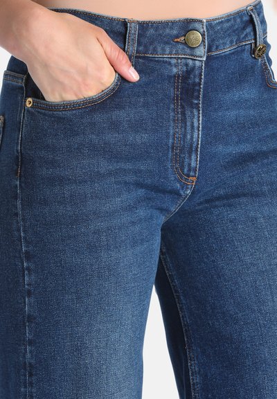 Main reposant dans la poche avant d'un jean en denim bleu avec un bouton en laiton visible et des détails de couture.