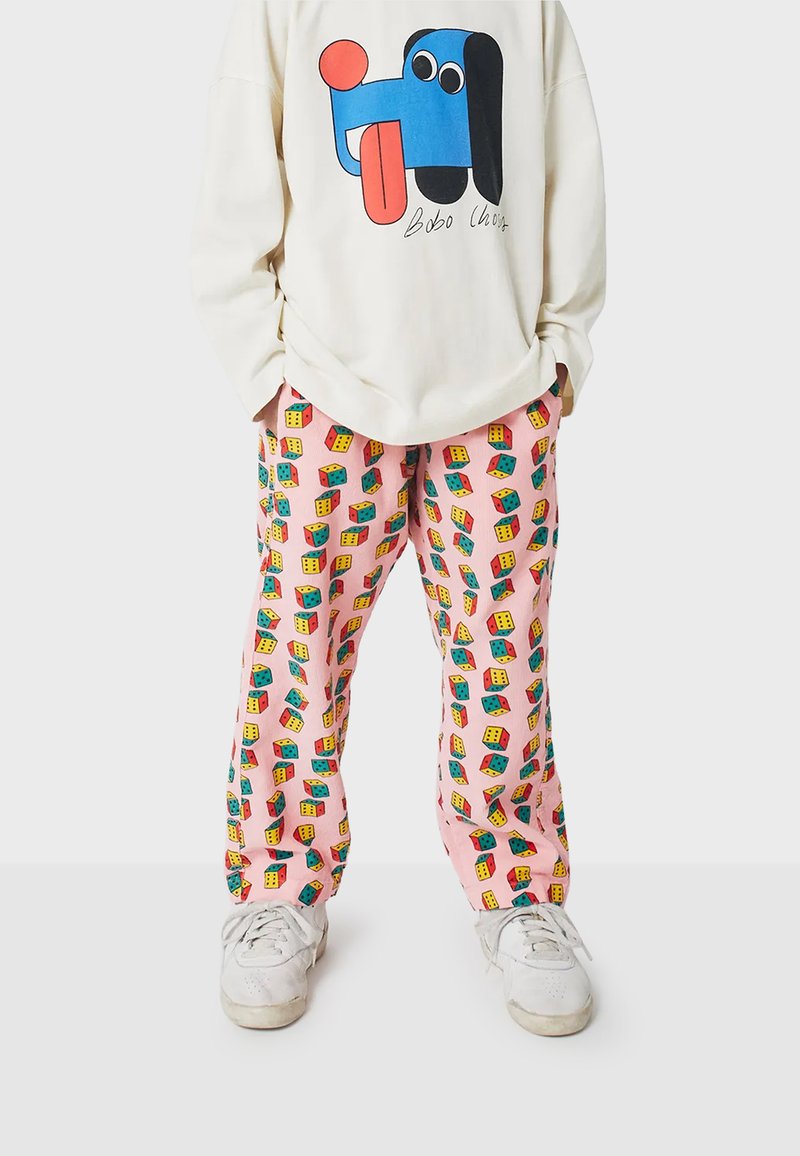 Pantalon de survêtement rose avec des motifs de cubes colorés, associé à un sweat-shirt graphique beige arborant un motif de chien bleu et des baskets blanches.