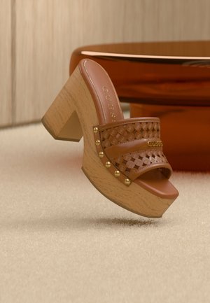 Mule à talon haut en cuir marron avec bride cloutée et découpée, talon compensé en bois, posé sur un tapis beige près d'un meuble orange-brun.