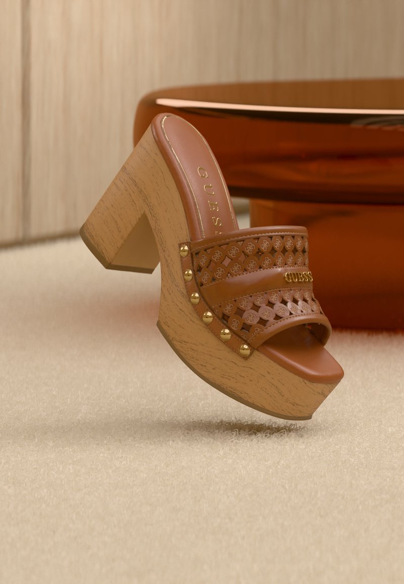 Καφέ δερμάτινο mule με ψηλό τακούνι, λουρί με καρφιά και κοψίματα, ξύλινο τακούνι πλατφόρμας, ακουμπά σε μπεζ χαλί κοντά σε πορτοκαλοκάστανα έπιπλα.