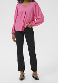 Blouse rose à col en V avec manches longues bouffantes, associée à un pantalon noir ajusté. La blouse présente un pli décoratif et des poignets froncés.