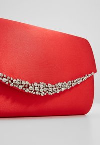 Pochette en satin rouge avec une rangée courbée de strass clairs scintillants sur le rabat avant sur un fond neutre.