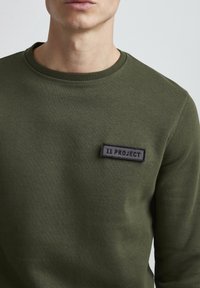 Sudadera verde oliva de algodón, con cuello redondo y un parche de etiqueta negra con "11 PROJECT" bordado en el pecho.