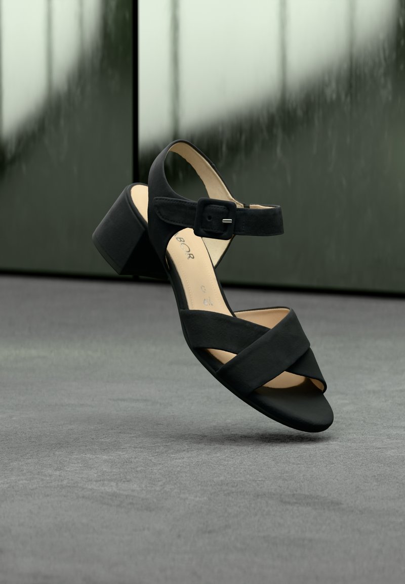 Μαύρο σανδάλι με ψηλό τακούνι και σταυρωτά λουριά, αγκράφα στον αστράγαλο, λεία υφή και γερό τακούνι σε σχήμα μπλοκ. Η σόλα έχει ανοιχτόχρωμο χρώμα.