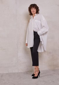 Kvinna med lockigt hår iklädd en överdimensionerad vit skjorta med knappar, svarta cropped byxor och svarta klackar med gyllene ankelkedjor, stående mot en vägg.