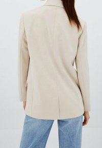 Beige takattu bleiseri, jossa on jämäkkä muotoilu, pitkät hihat ja halkio takana, yhdistettynä vaaleansinisiin farkkuihin.