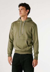 Man in een olijfgroen hoodie met een voorzak en witte trekkoorden, staande tegen een effen lichte achtergrond, kijkend naar de zijkant.