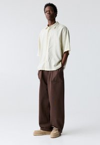 Chemise boutonnée à manches courtes de couleur jaune clair avec des rayures verticales, associée à un pantalon large marron large et des chaussures beige.