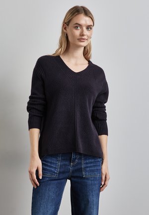 Donkerblauwe gebreide trui met V-hals, geribbelde textuur, verlaagde schouders en een relaxte pasvorm, gecombineerd met blauwe jeans.