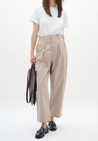 Beige kalhoty s vysokým pasem, sklady, zkombinované s bílým tričkem a černými koženými mokasíny, držící hnědou koženou tašku. Hladký materiál, volný střih.