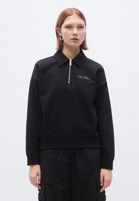 Felpa nera con collo con zip e polsini a costine. Presenta un logo ricamato sul petto e una vestibilità ampia. Tessuto strutturato.