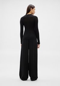 Sieviete redzama no muguras, valkājot pieguļošu melnu ribotu garroku džemperi un platu melnu bikses ar plašām kājām, uz vienkrāsaina gaiša fona.