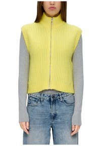 Gelber gestrickter ärmelloser Pullover mit hohem Kragen und Reißverschlussdetails, kombiniert mit einem grauen Langarmshirt unter blauen Jeans.