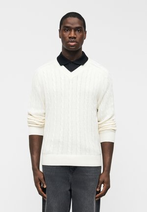 Mężczyzna ubrany w biały sweter warkoczowy z dekoltem w serek, nałożony na czarną koszulę z kołnierzykiem oraz ciemnoszare spodnie, stojący na jednolitym tle.