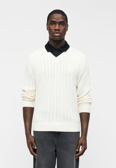 Homme portant un pull blanc en maille torsadée à col en V sur une chemise noire à col et un pantalon gris foncé, debout devant un fond uni.