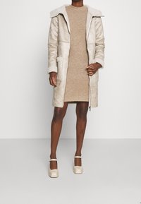 Cappotto di shearling beige con grandi tasche e cappuccio, indossato su un vestito di maglia marrone chiaro. Scarpe beige con tacco a blocco e cinturini alla caviglia.