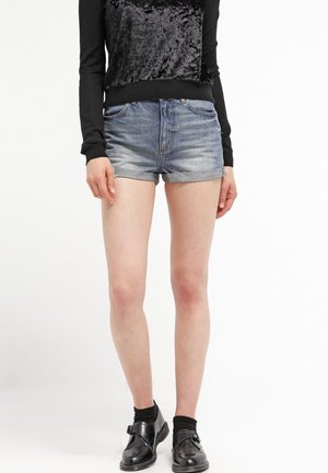 Shorts de denim en azul desgastado con dobladillo enrollado, acompañados de una blusa negra de manga larga con un panel de terciopelo en el frente.