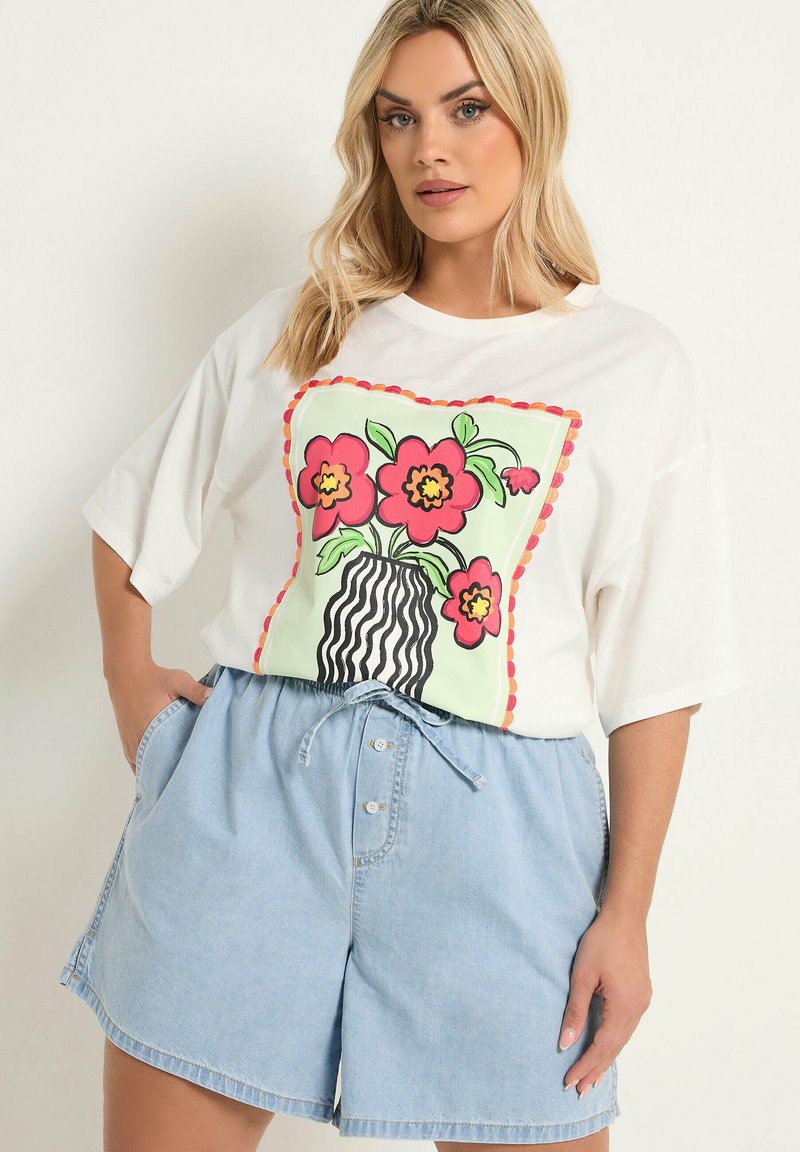 Donna che indossa una t-shirt bianca oversize con grafica di fiore rosso e vaso a righe, abbinata a pantaloncini di jeans azzurri a vita alta.
