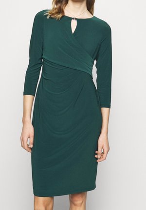 Frau trägt ein dunkelgrünes knielanges Kleid mit dreiviertel Ärmeln und einem Wickel-Stil Oberteil mit kleinem Schlüsselloch-Ausschnitt.