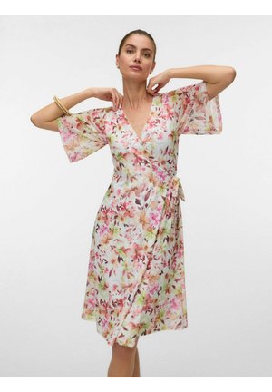 Naine, kes kannab valget põlveni olevat mähisrõivast roosa ja rohelise lillemustriga, poseerib käed kaela ja vöökoha lähedal.