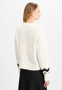 Cremfarbener, gestrickter Pullover mit einer weichen, flauschigen Textur, ausgestattet mit gerippten Bündchen und einem schwarzen Zickzackmuster an den Ärmeln.