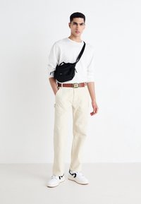 Bílá košile s dlouhým rukávem, béžové kalhoty, černá taška přes rameno, hnědý opasek se stříbrným akcentem, bílé tenisky s černými detaily.