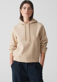 Felpa beige con cappuccio e cordino, dalla texture morbida, con polsini e orlo a costine. Presenta un dettaglio logo discreto e una silhouette dal taglio rilassato.