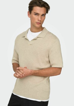 Ung mann med stylet brunt hår iført en beige kortermet genser med krage over en hvit skjorte, hendene foldet, mot en ensfarget bakgrunn.