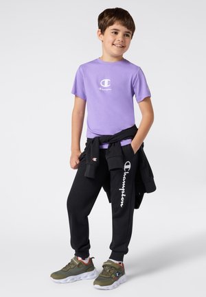 Lila T-Shirt mit kurzen Ärmeln und weißem Logo, schwarze Jogginghose mit Logo-Print, grüne Turnschuhe mit Akzenten, getragen mit einem schwarzen Sweatshirt, das um die Taille gebunden ist.