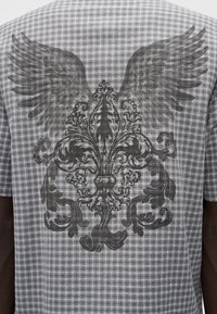 Γκρι πουκάμισο με κοντά μανίκια και καρώ σχέδιο, με μεγάλο μαύρο γράφημα με φτερά και περίτεχνη διακόσμηση στην πλάτη, υφασμένο με υφή.