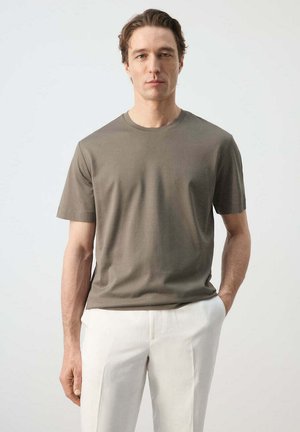 Bărbat purtând un tricou verde măsliniu cu mâneci scurte și pantaloni albi, stând cu o mână în buzunar, pe un fundal simplu, deschis la culoare.